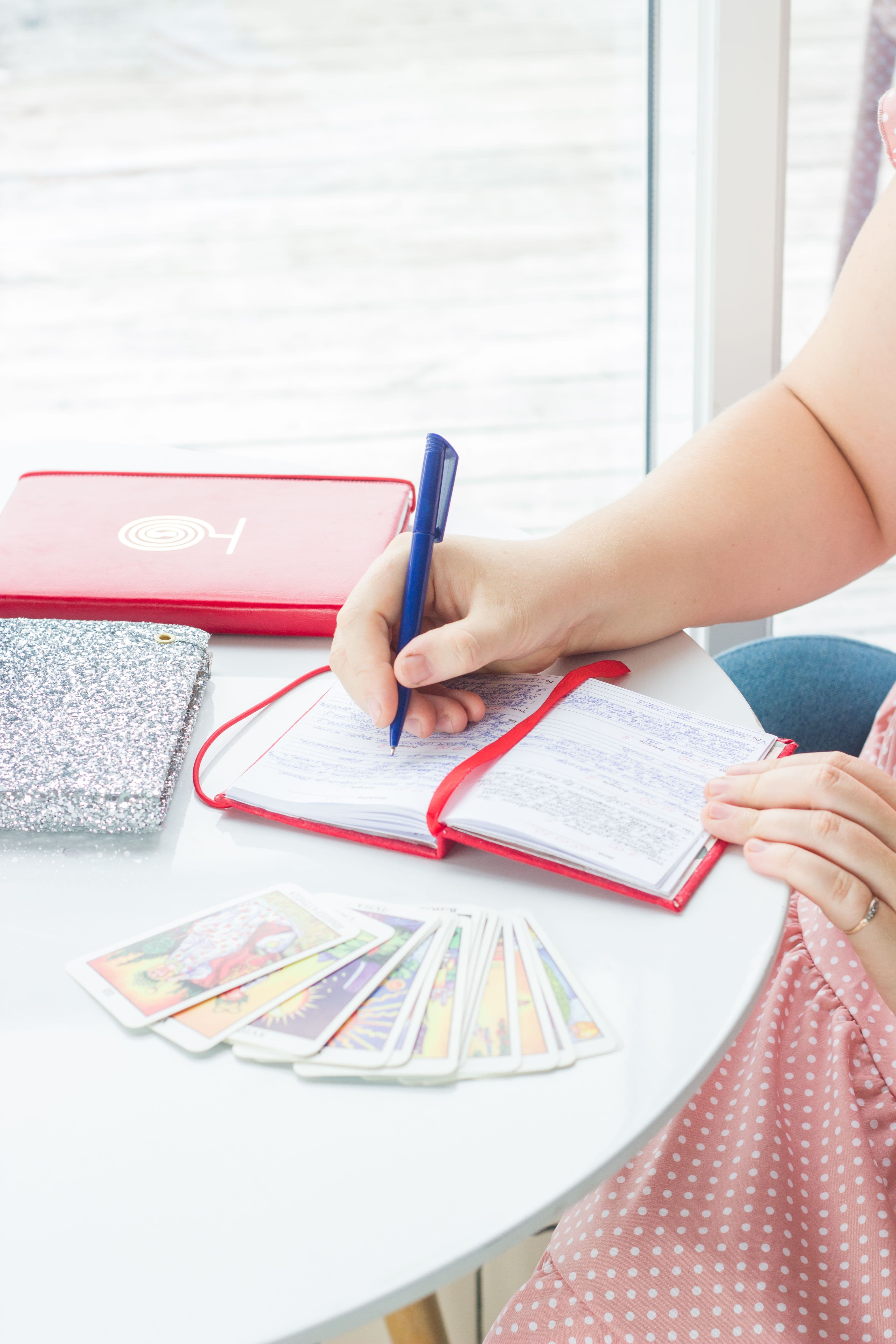 Woman writing in a red notebook with tarot cards fanned out on a white table.